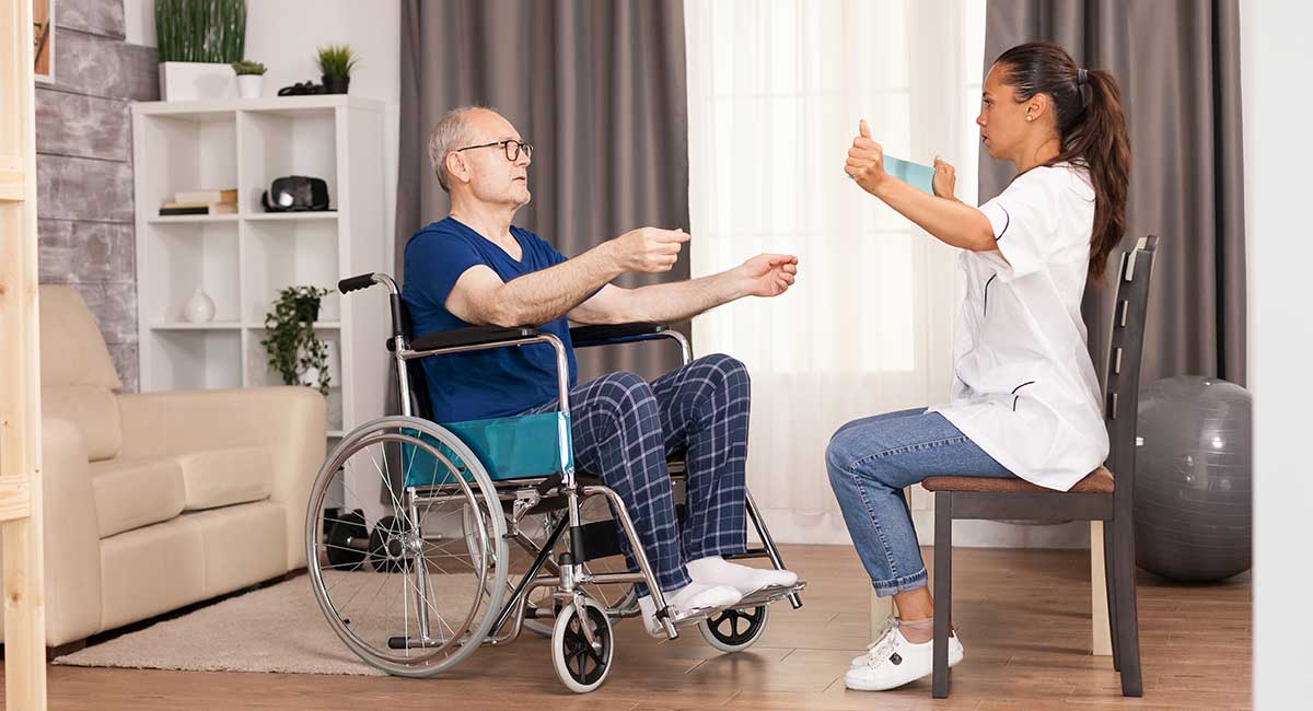 An in-home carer shows an elderly person in a wheelchair how to exercise.