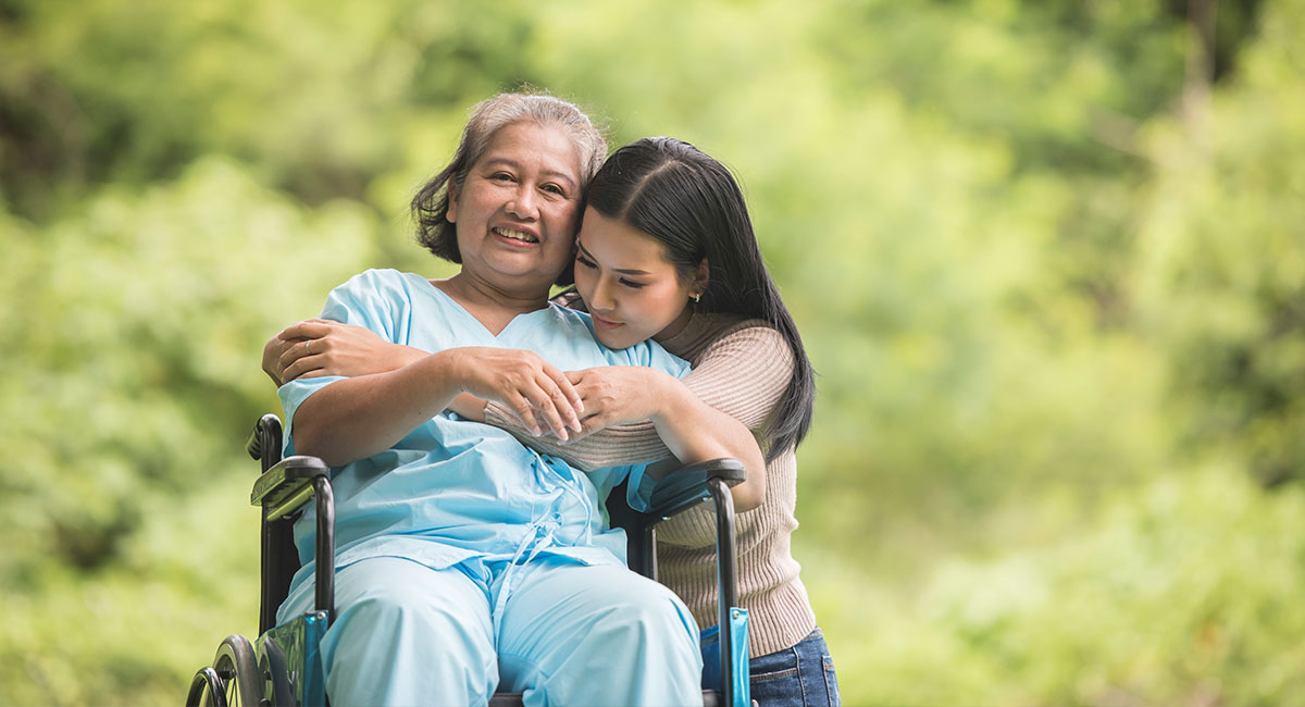 An elderly caretaker hugging a wheelchair-bound woman